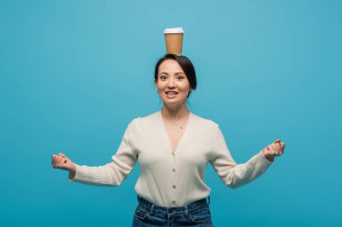 Excited asian woman with paper cup on head showing yes gesture isolated on blue 