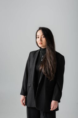Young asian woman in suit and turtleneck looking at camera isolated on grey 