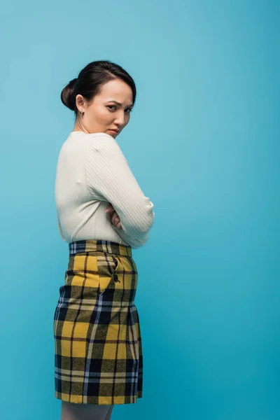 Embarrassed asian woman in skirt and cardigan looking at camera isolated on blue
