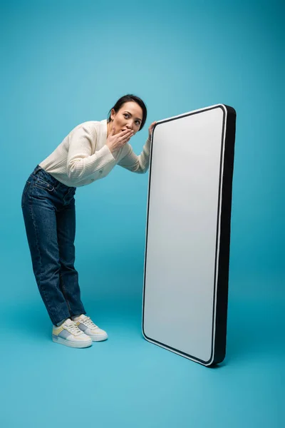 stock image Shocked asian woman covering mouth near big smartphone model on blue background