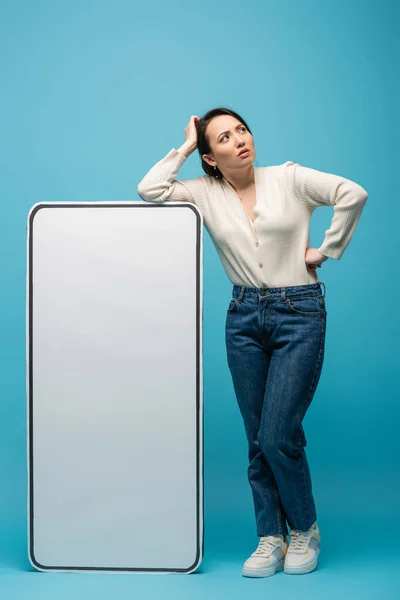 stock image Full length of pensive asian woman standing near big smartphone mockup on blue background