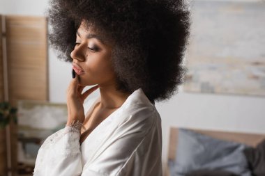 Beyaz saten bornoz giymiş düşünceli Afro-Amerikalı kadın evde dururken dudağına dokunuyor.