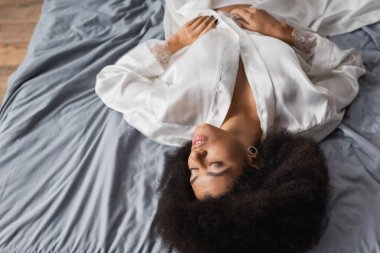 Esmer, Afro-Amerikan kadının yüksek açılı görüntüsü. Gözleri kapalı, beyaz bornoz içinde gri yatak örtüsünün üzerinde yatıyor.
