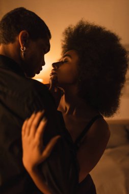 Baştan çıkarıcı Afro-Amerikalı kadın, gözleri kapalı, karanlık odada ışıkla genç adama sarılıyor.