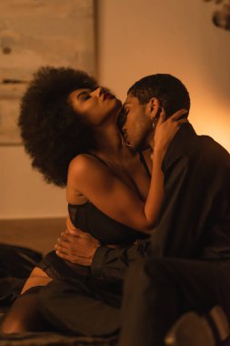 side view of african american man hugging and kissing seductive girlfriend in bedroom at night clipart