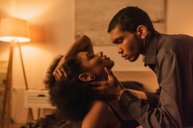 Siyah tişörtlü Afro-Amerikan bir adam yatak odasında baştan çıkarıcı kız arkadaşının boynuna ışıkla sarılıyor.