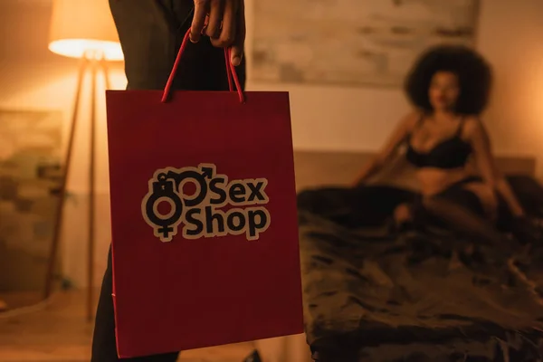 stock image partial view of young man holding paper bag from sex shop near blurred african american on bed at home