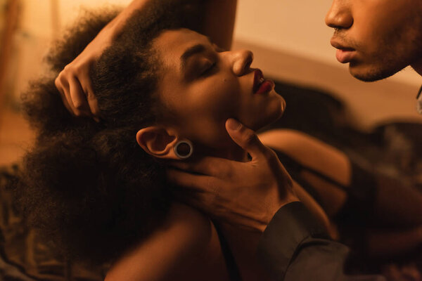 young man touching neck of seductive african american girlfriend at night at home
