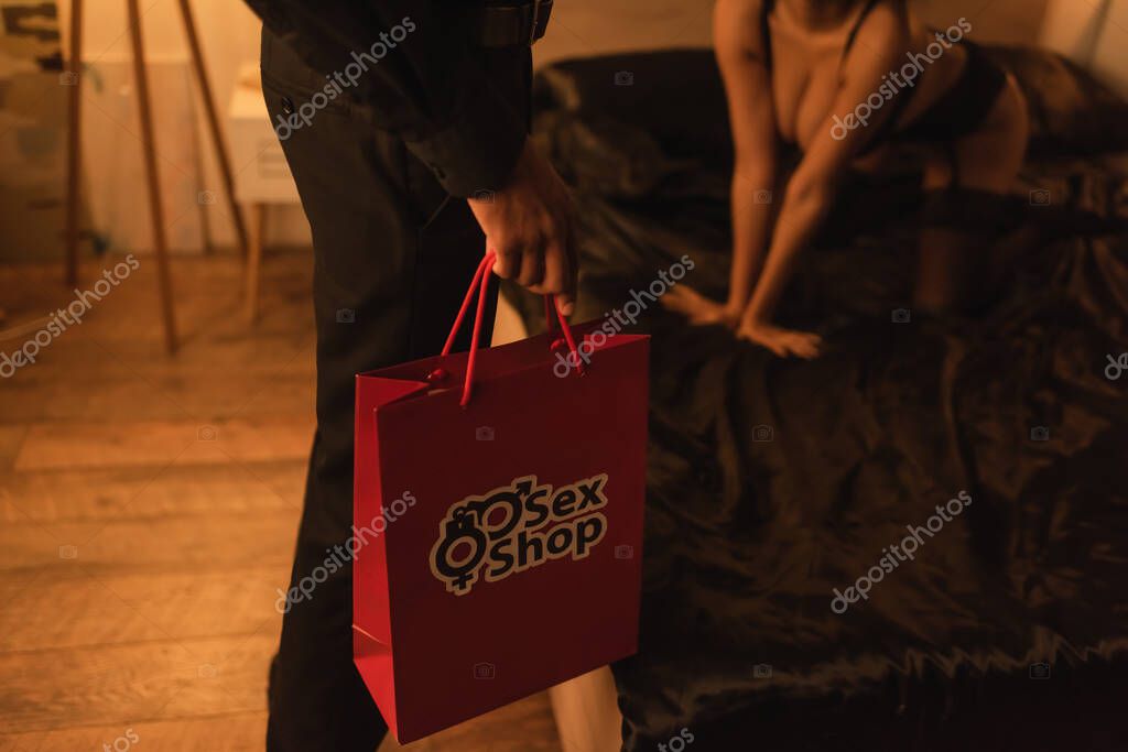Cropped view of man in black clothes holding shopping bag with sex shop lettering near sexy african american woman on bed