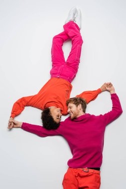 top view of happy redhead man and curly african american woman in magenta color clothes holding hands on white  clipart