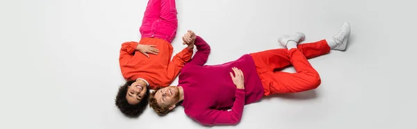 Stock image top view of happy redhead man and joyful african american woman in magenta color clothes lying on white, banner 