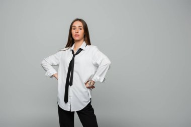confident young woman in white shirt and tie looking at camera while standing with hands on hips isolated on grey  clipart
