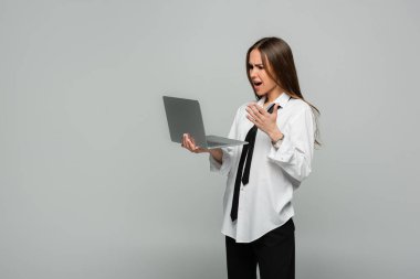 displeased young woman in white shirt with tie using laptop isolated on grey  clipart