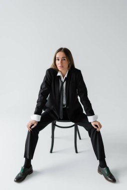 full length of young brunette woman in black suit with tie sitting on wooden chair on grey 