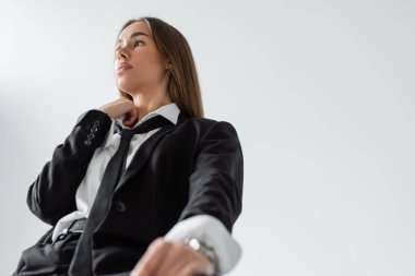 low angle view of young brunette woman in black suit with tie sitting and looking away on grey 
