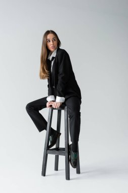 full length of young brunette woman in black suit with tie sitting on high chair and pouting lips on grey 