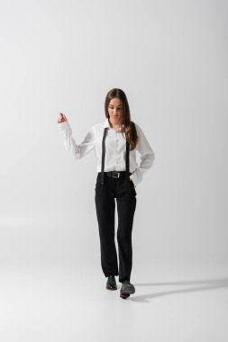 full length of brunette woman in black pants and suspenders walking with hand in pocket on grey  clipart