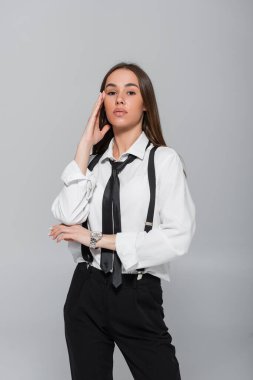brunette young woman in black pants with suspenders looking at camera isolated on grey 