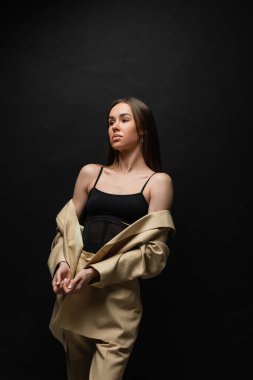 brunette young woman in beige jacket and trousers standing and looking away on black 