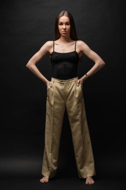 full length of muscular woman in top and beige pants posing with hands in pockets on black background  clipart