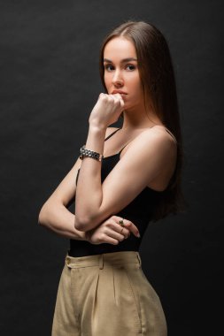 brunette woman in top and beige pants posing while looking at camera on black background 
