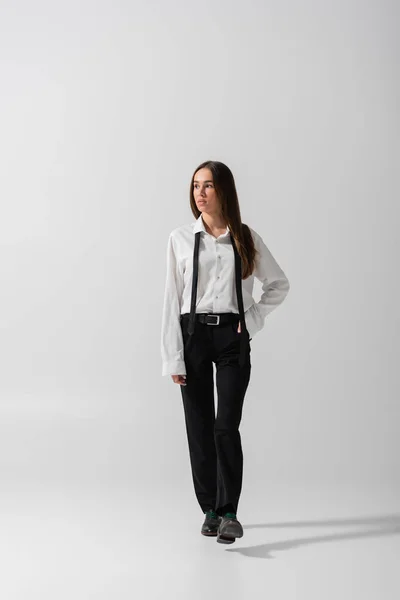 stock image full length of brunette woman in black pants and suspenders standing on grey 