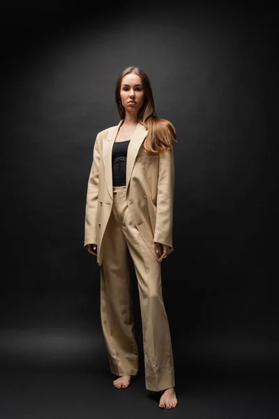 full length of barefoot woman in beige suit standing and looking at camera on black