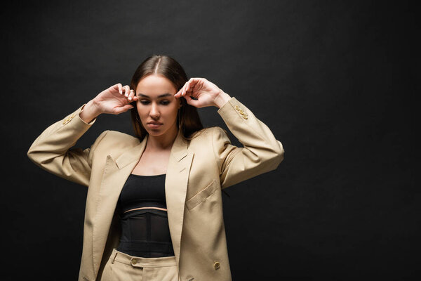 брюнетка молодая женщина в бежевом пиджаке стоя и глядя вниз на черном фоне 