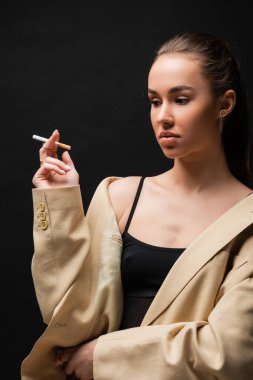 young brunette woman in beige blazer holding cigarette on black background 