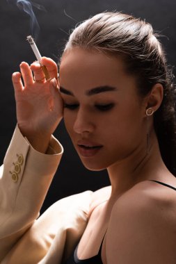 portrait of young brunette woman in beige blazer holding cigarette and looking away on black background  clipart