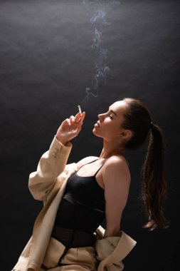 side view of young brunette woman in beige blazer holding cigarette on black background  clipart