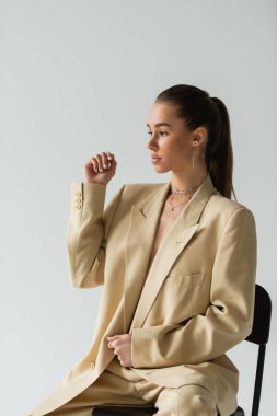 pretty young woman in beige trendy suit sitting on chair isolated on grey 