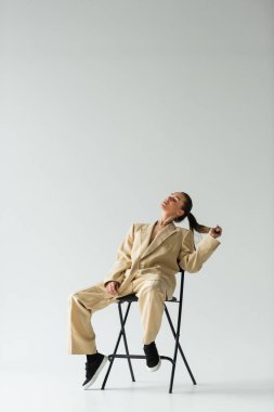 full length of young woman in beige trendy suit sitting on chair and adjusting ponytail isolated on grey 