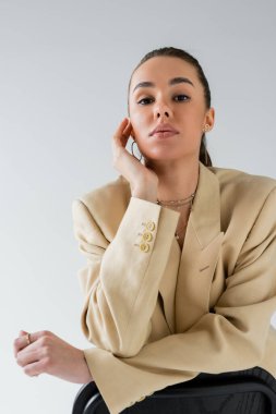 brunette young woman in beige jacket leaning on chair back while looking at camera isolated on grey 
