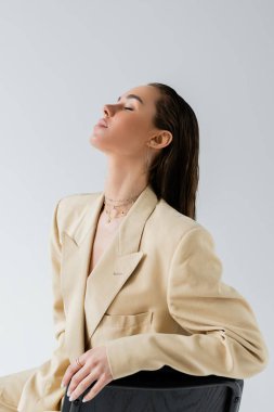 pretty young woman in beige jacket sitting on chair and posing with closed eyes isolated on grey 
