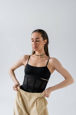 pretty model in black top standing with closed eyes and adjusting beige pants isolated on grey 