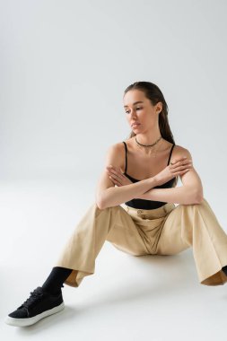 full length of thoughtful woman in beige pants and black top sitting on grey 