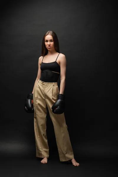 stock image full length of strong woman in top and beige pants posing in boxing gloves on black background 