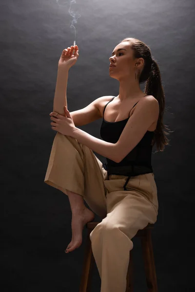 pensive woman in beige pants holding cigarette while sitting on high chair on black background