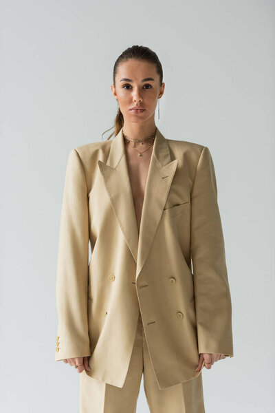 brunette young woman in beige jacket posing while looking at camera isolated on grey 
