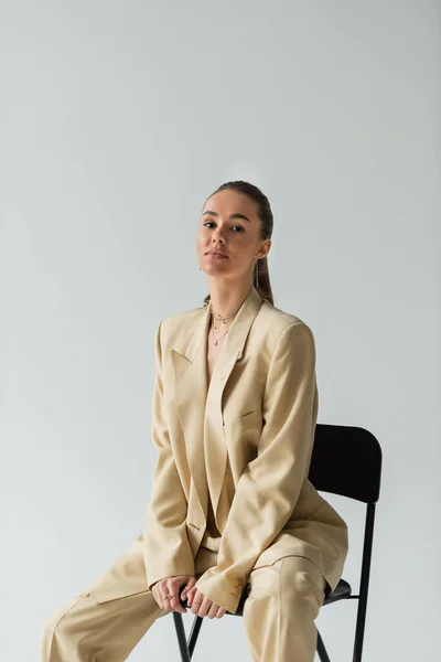 stock image brunette young woman in beige trendy suit sitting on chair isolated on grey 