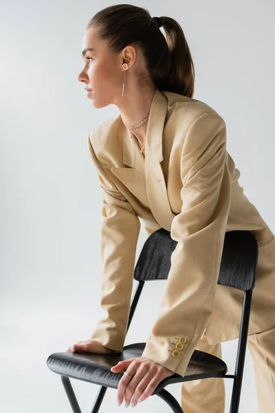 pretty young woman in beige jacket leaning on chair while looking away isolated on grey