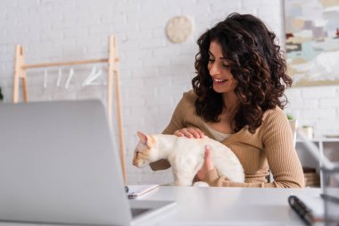 Positive freelancer looking at oriental cat near laptop and notebooks on table 