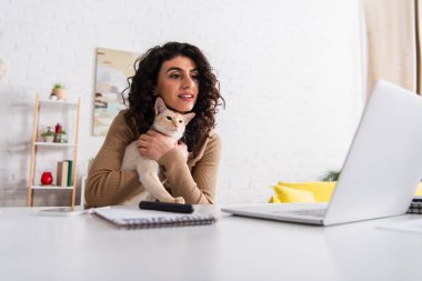 Smiling copywriter holding oriental cat and looking at laptop in living room 