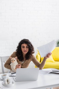 Copywriter holding oriental cat and papers near laptop at home 