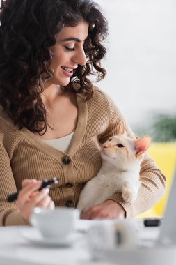 Smiling copywriter holding marker and looking at oriental cat near cup and laptop at home 