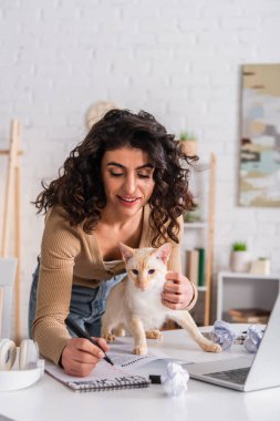 Cheerful copywriter writing on notebook near oriental cat and laptop 