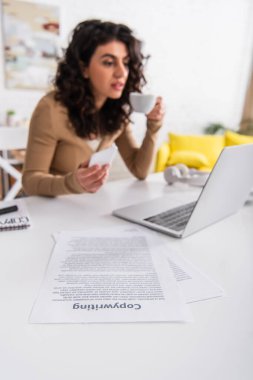 Papers with copywriting lettering near blurred woman holding cup and smartphone at home  clipart