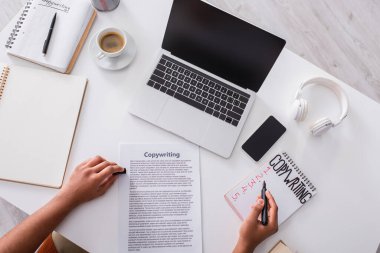 Top view of copywriter writing on notebook near devices with blank screen and coffee on table 