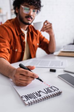 Blurred indian freelancer holding marker near notebook with copywriting lettering and coffee at home 